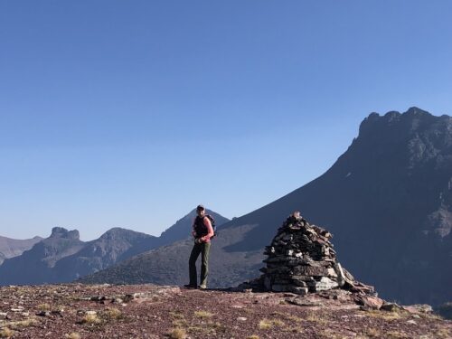 Woman wearing hiking gear standing next to a tall cairn on top of a mountain surrounded by peaks. 