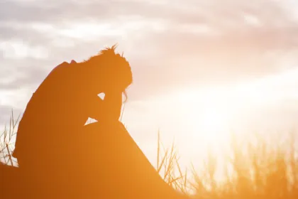 Person sitting on ground, legs bent and torso bent over with head in her heands like in despair with bright sky and clouds behind her, grass glowing golden.