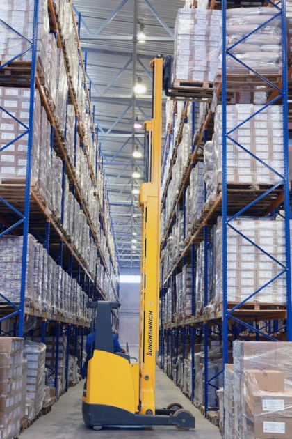 Person standing in a stand-up electric forklift in a warehouse between racks with pallets in them with forks raised sliding in or removing a pallet from the rack.