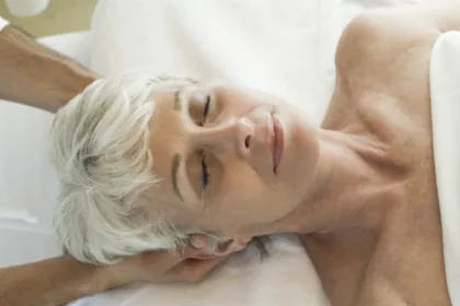 Older, white-haired woman lying on massage table with masseuses hands behind her head and her eyes closed, shoulders bare and sheet wrapped around her chest where picture ends.