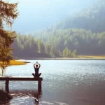 Woman sitting in yoga pose with hands together over her head on a dock that stretches out into a lake with a sunlit shore and forest behind it.