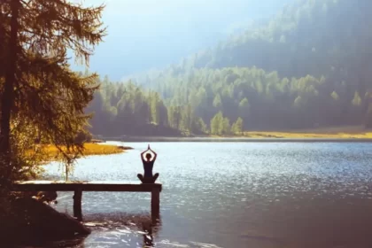 Woman sitting in yoga pose with hands together over her head on a dock that stretches out into a lake with a sunlit shore and forest behind it.