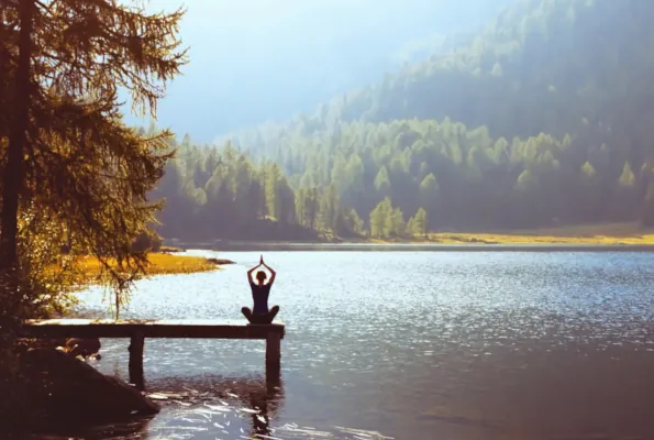 Woman sitting in yoga pose with hands together over her head on a dock that stretches out into a lake with a sunlit shore and forest behind it.