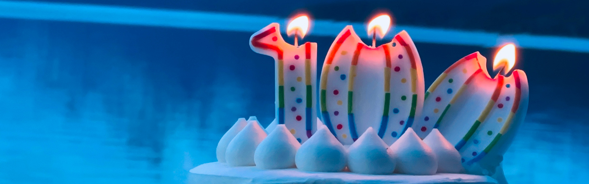 The number one hundred in three lit candles on top of a cake in front of a dark blue background.