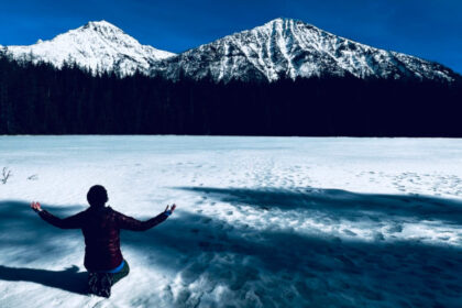 Bettina Theresia kneeling in the snow that has the shadows of trees stretching accross it, looking up at snow-covered mountains with her arms raised out to the sides.