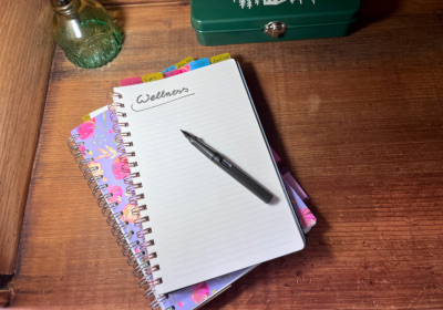 An opened lined notebook lying on a wooden desk with the word wellness written at the top of the page and an uncapped fountain pen lying on said page.