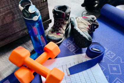 Grouping of a blue water bottle, two orange dumbbells, a yoga strap and some hiking boots on top of a yoga mat with a strength training log between them.