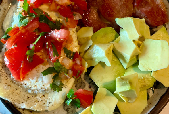 Fried eggs smothered with pico de gallo next to fried bacon and avocado pieces.